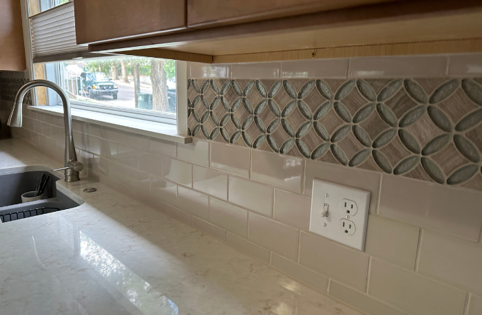 A kitchen counter with a sink and faucet, featuring a light-colored subway tile backsplash with a decorative mosaic tile border above it.